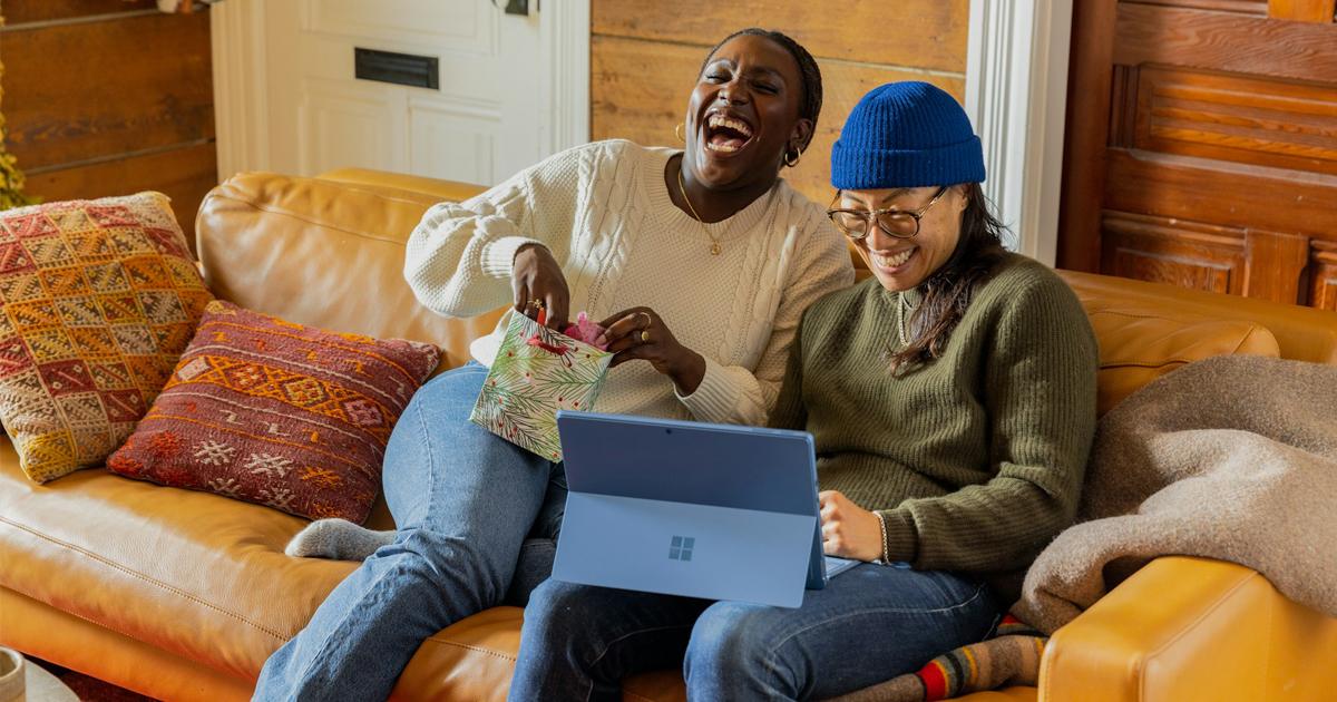 Two women sitting together on a leather sofa looking at a Microsoft Surface tablet while opening a gift bag.