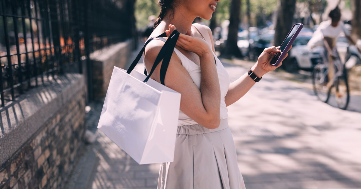 Young woman shopping outdoors on a sunny day, holding a white shopping bag and using a smartphone — lifestyle, urban fashion, and mobile commerce concept.