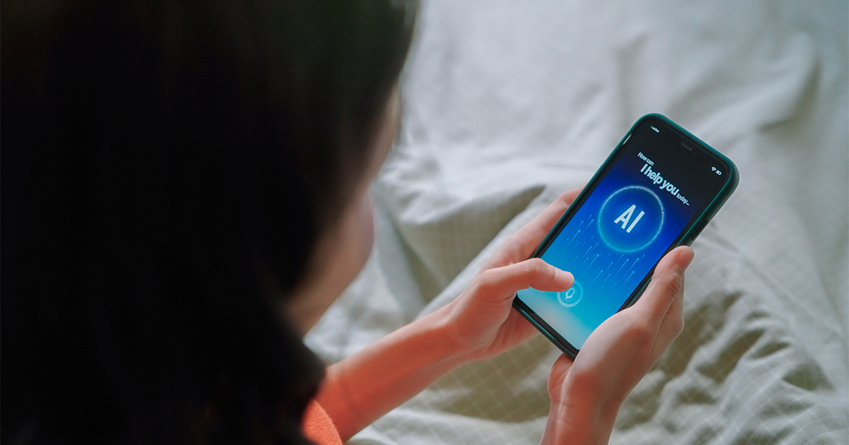 Person using a smartphone with an AI assistant interface displayed on the screen, sitting on a bed with a soft gray blanket.