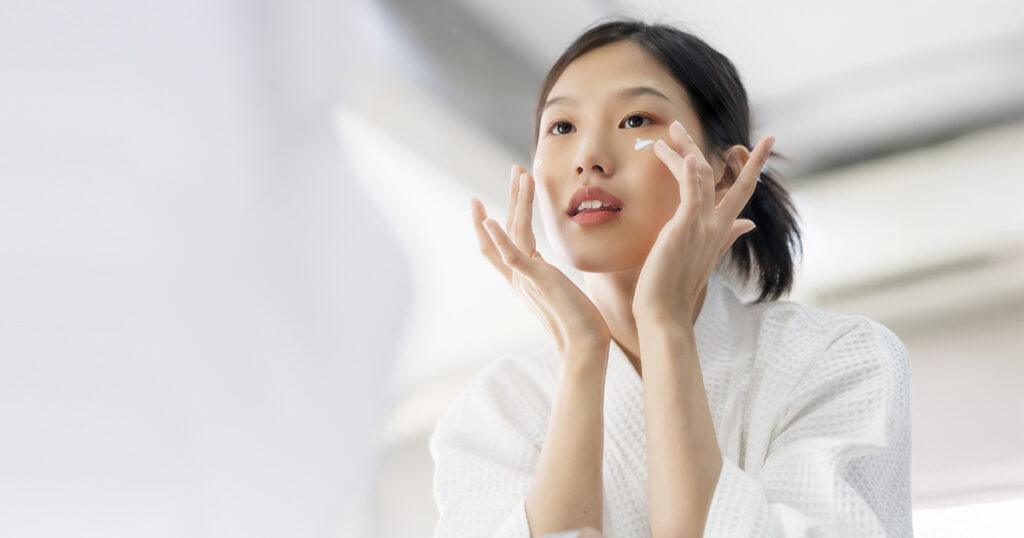A young woman with her hair pulled back, wearing a white robe, applies a cream to her face. She looks into a mirror, using her fingers to gently dab the cream below her eye.