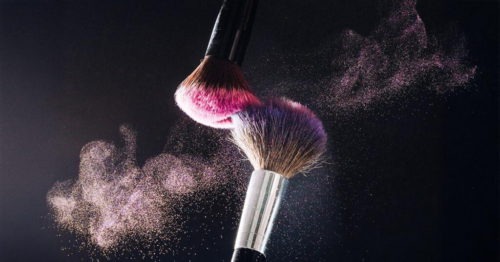 Two makeup brushes tap together, one with pink bristles and one with purple, creating a puff of shimmering pink powder that floats in the air against a dark background.