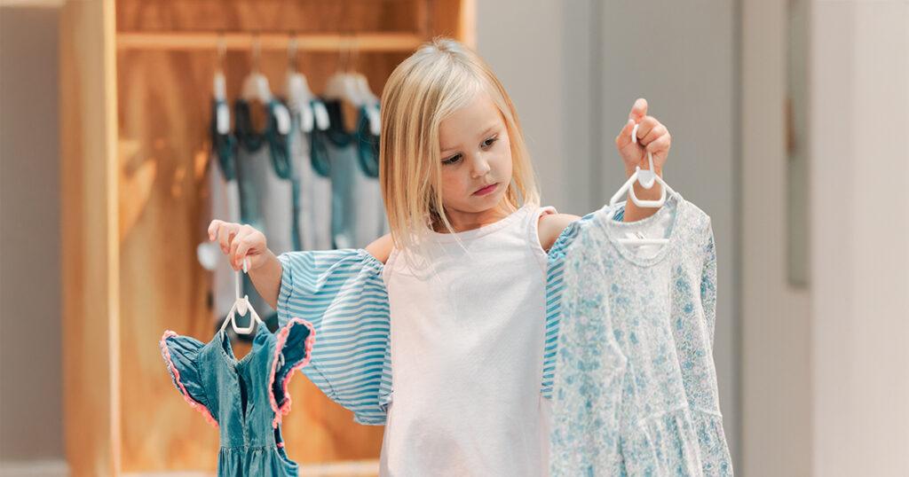 A blonde young Gen Alpha girl stands in a retail store comparing two dresses symbolizing the consumer influence of the next generation of shoppers.