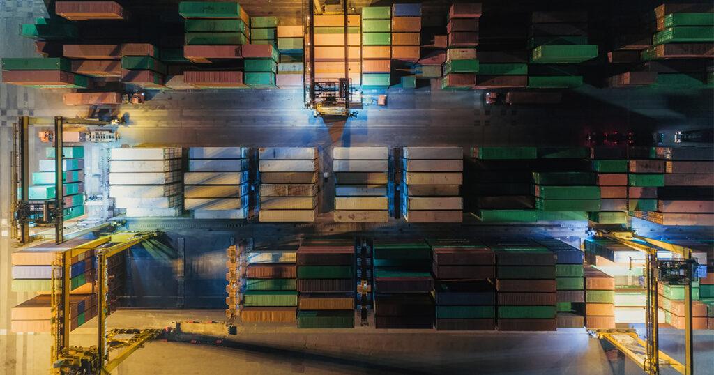 Aerial view of a shipping port at night with rows of stacked cargo containers and cranes.