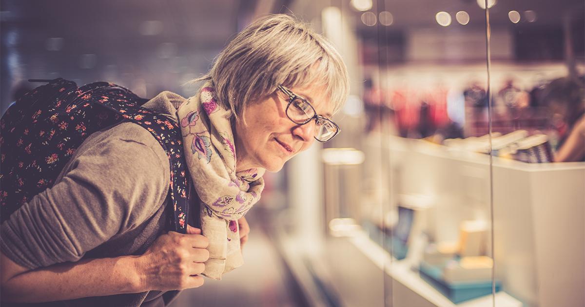 A middle-aged woman with short gray hair, glasses, a floral scarf, and a backpack leans forward with curiosity to look through a shop window.