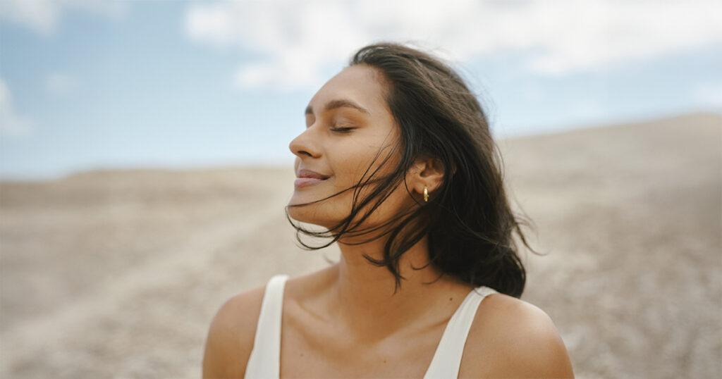 Young Indian woman outdoors with eyes closed, enjoying the breeze in a natural landscape under a blue sky.