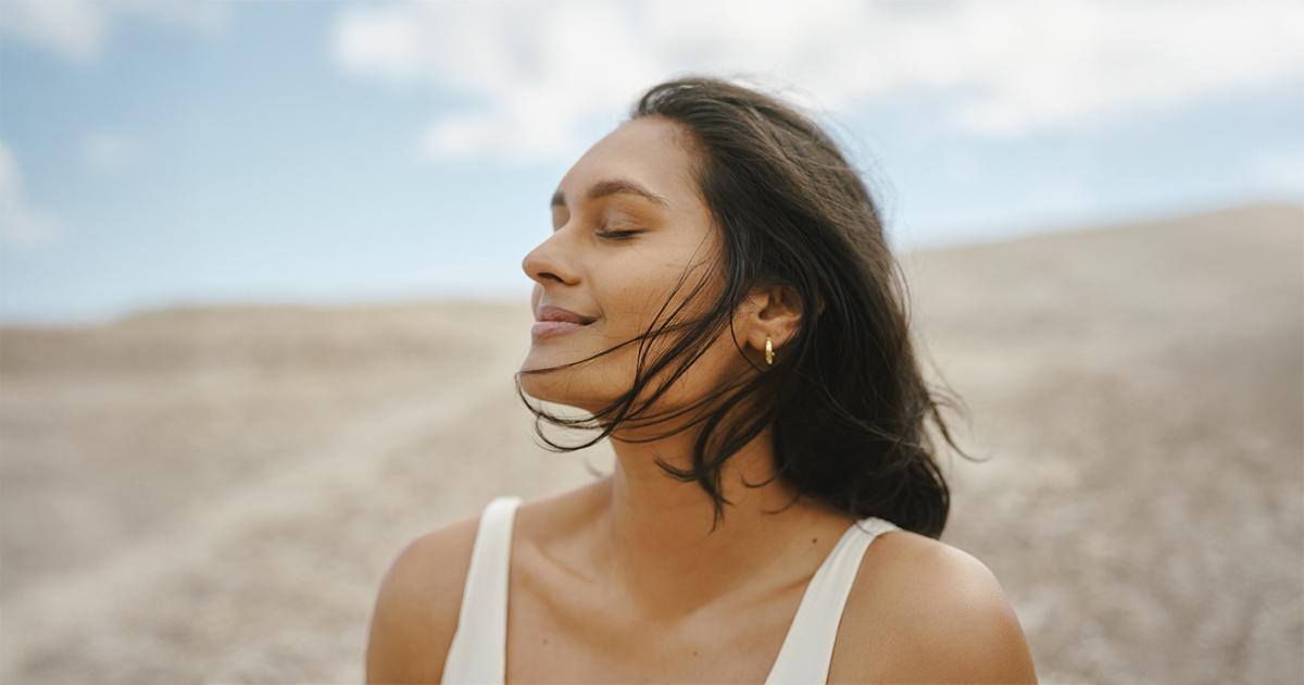 Young Indian woman outdoors with eyes closed, enjoying the breeze in a natural landscape under a blue sky.