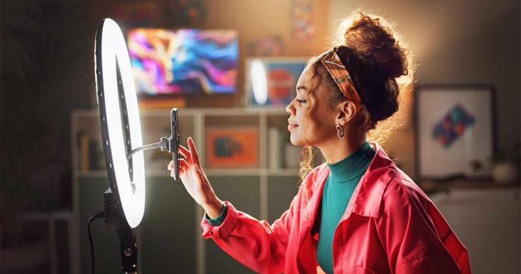 Young woman recording content on her smartphone with a ring light, wearing a bright pink jacket and headband at home.