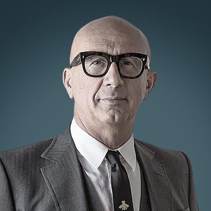 Marco Bizzarri, Co-Founder and President of Forel, wearing a grey suit and black glasses against a blue background.