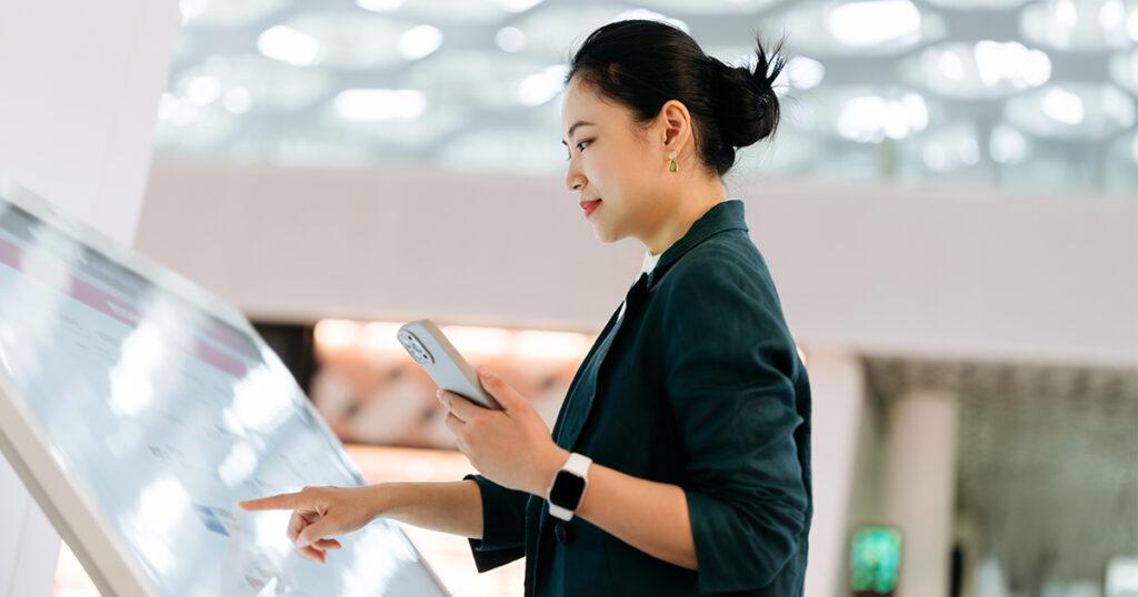An Asian woman using a digital touchscreen kiosk while holding a smartphone in a bright indoor setting