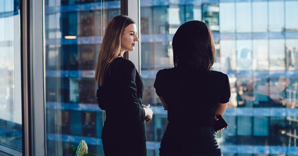 Two women in business attire standing before a large office window, looking out at modern glass buildings in the city.