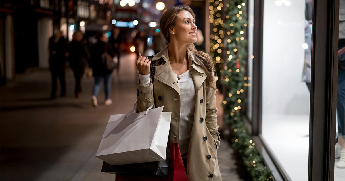 A woman in a beige trench coat and carrying shopping bags is smiling while looking at a store window with Christmas decorations.