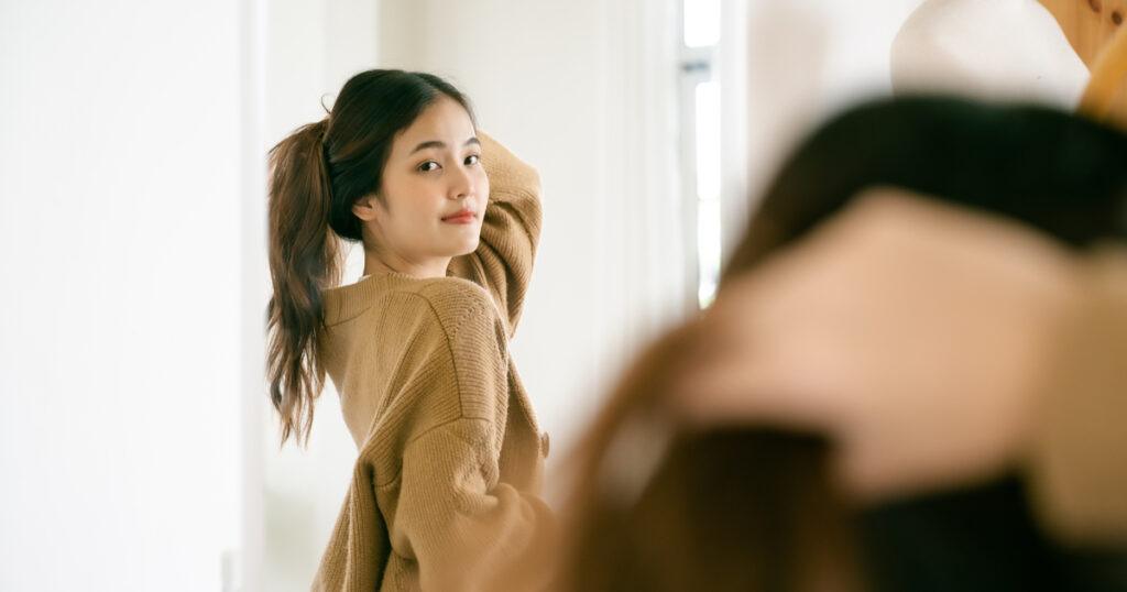 A young Asian woman in a brown sweater looks over her shoulder, holding her long hair and smiling slightly in front of a mirror.
