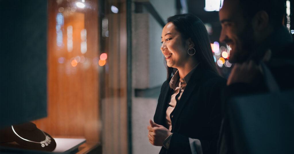 A smiling woman and man outside a jewelry store at night, looking through the window display at a necklace.