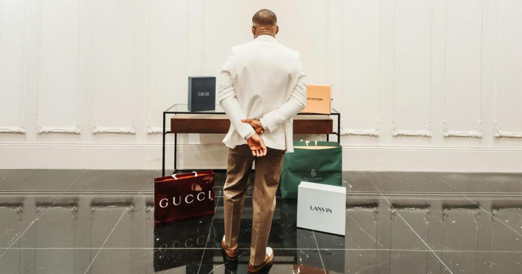 A man in a white blazer stands in a room, facing luxury shopping bags from Gucci, Dior, Louis Vuitton, Goyard, and Lanvin displayed on the floor.