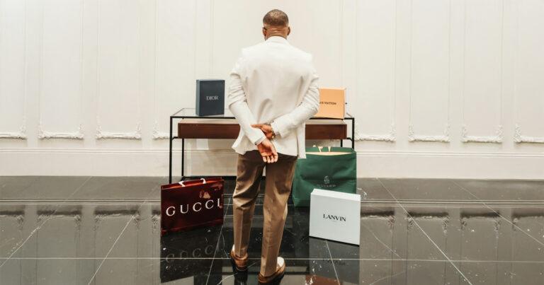 A man in a white blazer stands in a room, facing luxury shopping bags from Gucci, Dior, Louis Vuitton, Goyard, and Lanvin displayed on the floor.