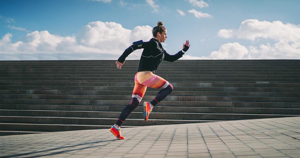 A woman wearing colorful running tights and a black top and listening to music on her phone runs up an outdoor staircase.