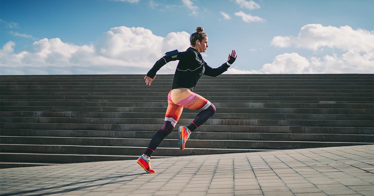 A woman wearing colorful running tights and a black top and listening to music on her phone runs up an outdoor staircase.