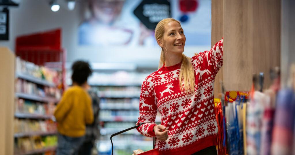 A blonde woman wearing a red Christmas sweater smiles as she reaches for an item on a grocery store shelf, with other shoppers visible in the background among the aisles.