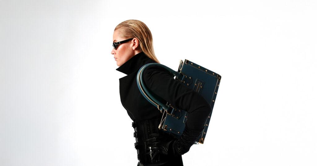 A blonde model wearing a high-collared black coat, black sunglasses and black gloves carries a trunk-style black handbag over her shoulder against a white background.