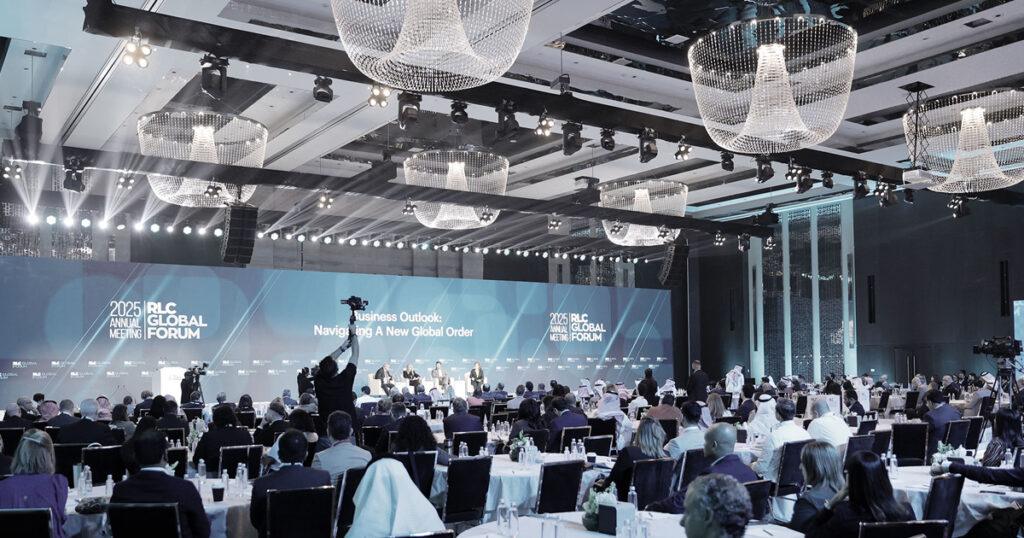 A large conference ballroom filled with attendees facing a stage with a panel of speakers. at the RLC 2025 Annual Forum. Bright stage lighting and multiple crystal chandeliers hang from the ceiling.
