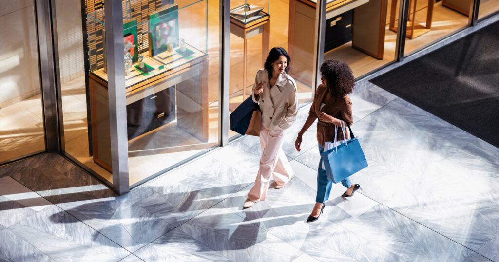 Two women walking through a shopping mall, carrying shopping bags and chatting in front of a luxury boutique with watches displayed in the window.