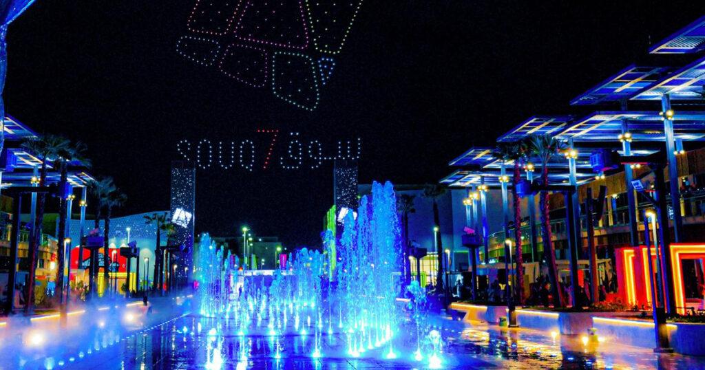 Nighttime view of Souq7 in Jeddah featuring illuminated fountains, vibrant lights and a drone show forming the Souq7 logo in the sky.