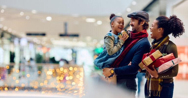 A smiling family is carrying Christmas gifts while shopping in a shopping mall decorated with festive lights.