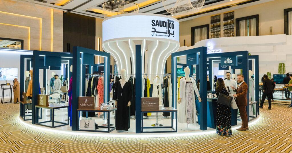 Exhibition space showcasing Saudi 100 Brands, with fashion displays and mannequins arranged around a central illuminated structure.