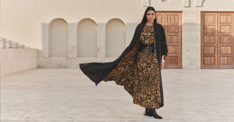 A model wearing a flowing leopard-print abaya with a black belt and a long black cape stands in front of a building with arched windows and wooden doors.