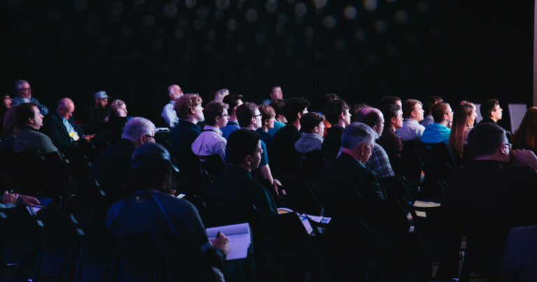 Audience seated in a dark auditorium watching a presentation or performance, faces lit by stage light.