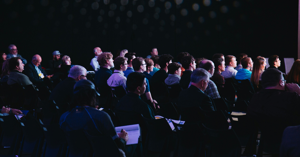 Audience seated in a dark auditorium watching a presentation or performance, faces lit by stage light.