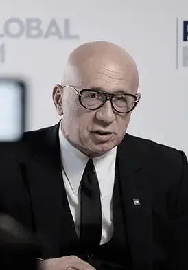 Man in a black suit and tie wearing glasses speaks at a conference or panel event.