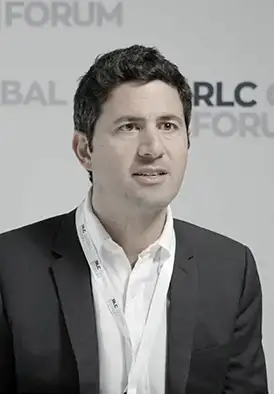 Man in a black suit and white shirt speaking at a forum event, with blurred forum branding in the background.