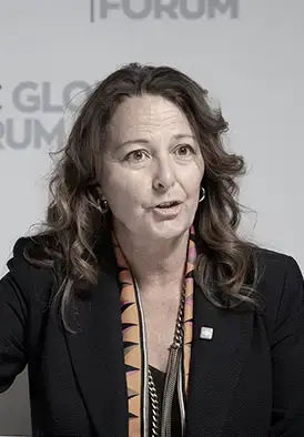 Woman with wavy brown hair speaking at a panel, wearing a black blazer and patterned scarf, in front of a Global Forum backdrop.