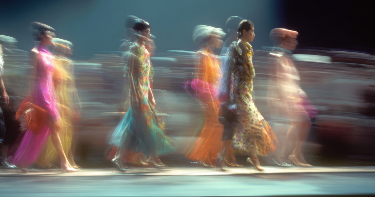 Blurred models in colorful dresses walk down a brightly lit runway.