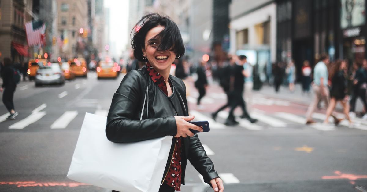 Smiling stylish woman holding a shopping bag and smartphone while crossing a busy street in New York City, surrounded by yellow taxis and pedestrians—symbolizing 2025 consumer behavior, shopping trends.