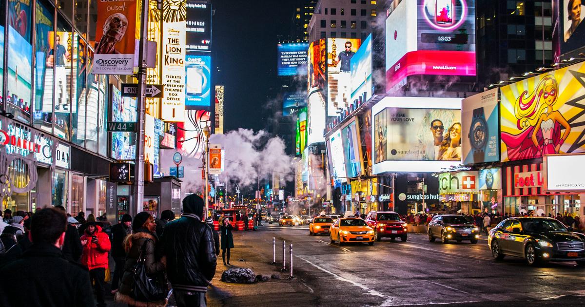A bustling Times Square street scene at night featuring illuminated digital billboards, retail storefronts, yellow taxis, and pedestrians.