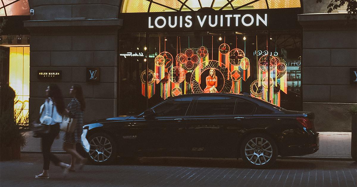 Two women walk past a Louis Vuitton storefront at dusk, with a luxury car parked in front. The window display features Louis Vuitton’s iconic geometric monogram motifs and a central mannequin, reflecting the blend of heritage and digital innovation and capturing the challenge luxury brands face in balancing tradition with AI-driven transformation.