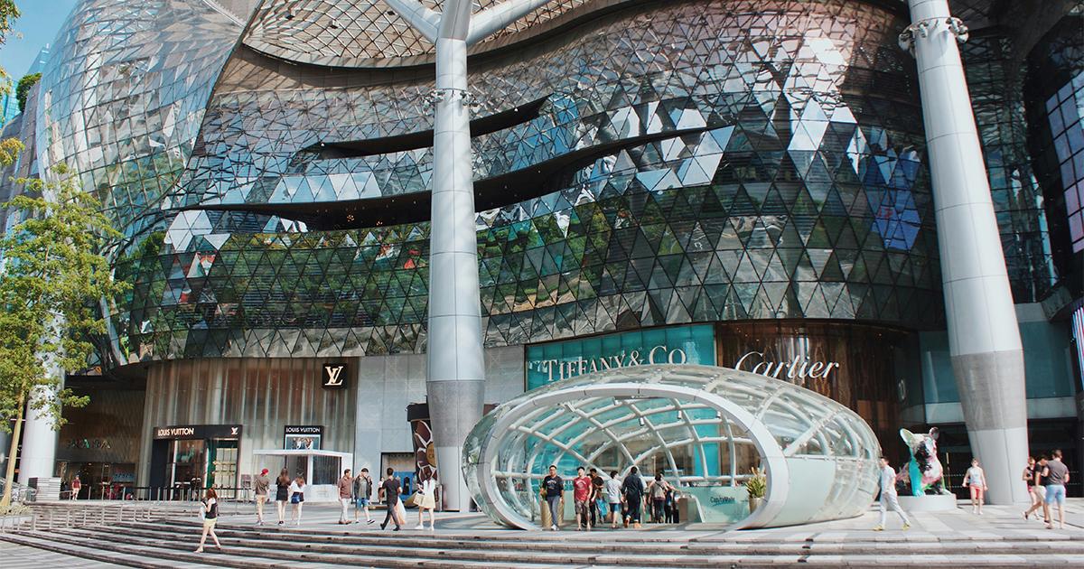 Futuristic glass retail building facade featuring luxury storefronts and a transparent oval entrance canopy at RLC Global Forum.