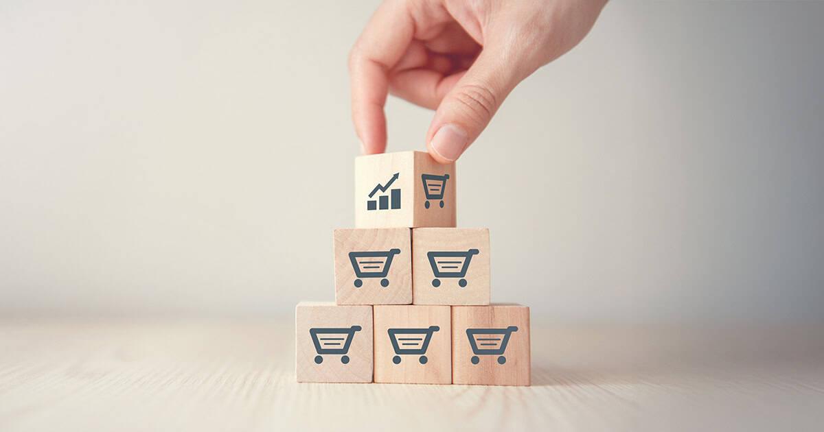 A hand placing a wooden block showing an upward growth chart icon atop a pyramid of blocks printed with shopping carts at RLC Global Forum.