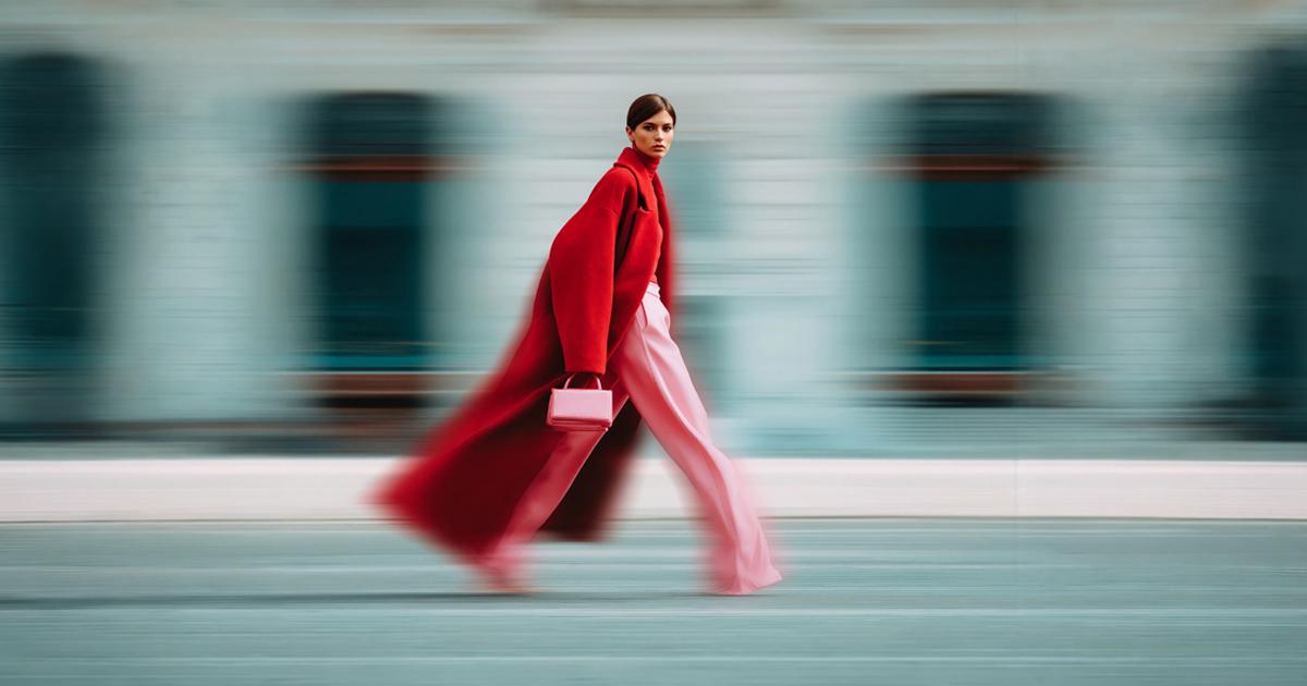 Young woman in a red coat and pink trousers holding a handbag while walking against a motion-blurred background.