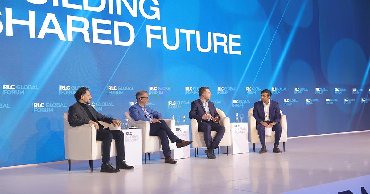 Four male speakers participating in a stage panel discussion in front of a blue RLC Global Forum backdrop.