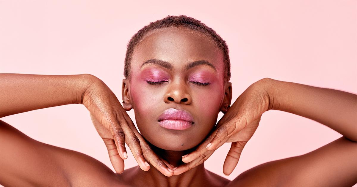 Portrait of a black woman with short hair and closed eyes, wearing pink eyeshadow and lipstick, against a pink background.