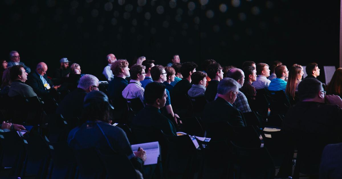 Audience seated in a dark auditorium watching a presentation or performance, faces lit by stage light.