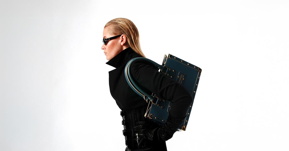A blonde model wearing a high-collared black coat, black sunglasses and black gloves carries a trunk-style black handbag over her shoulder against a white background.