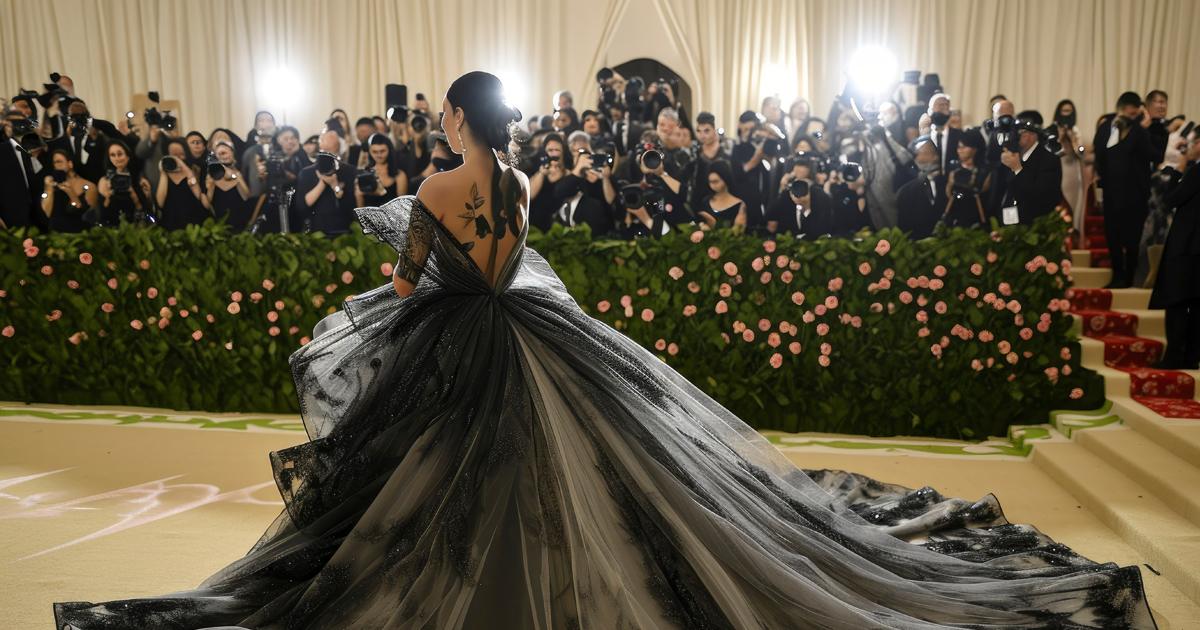 A woman in an ombre black gown poses on a carpeted staircase as photographers take pictures behind a floral hedge.