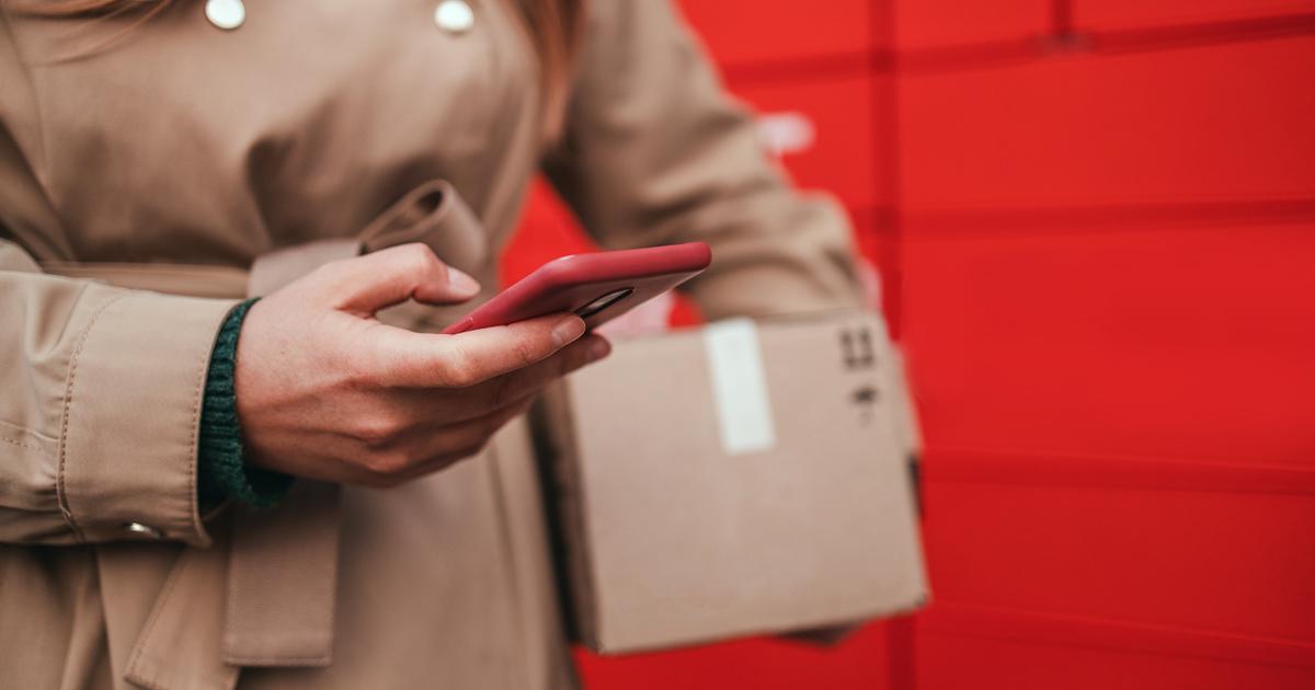 Person wearing a trench coat holds a smartphone and delivery parcel against a red locker background.