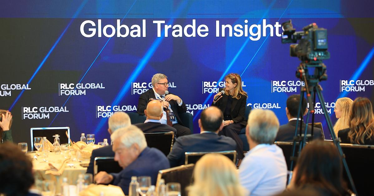 Two speakers on stage holding microphones during a Global Trade Insights panel at RLC Global Forum, viewed from behind the audience and a broadcast video camera.