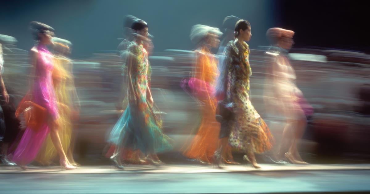 Blurred models in colorful dresses walk down a brightly lit runway.