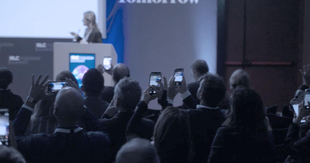 Audience members at the RLC Fashion Summit in Milan raise their phones to photograph a speaker presenting on stage.